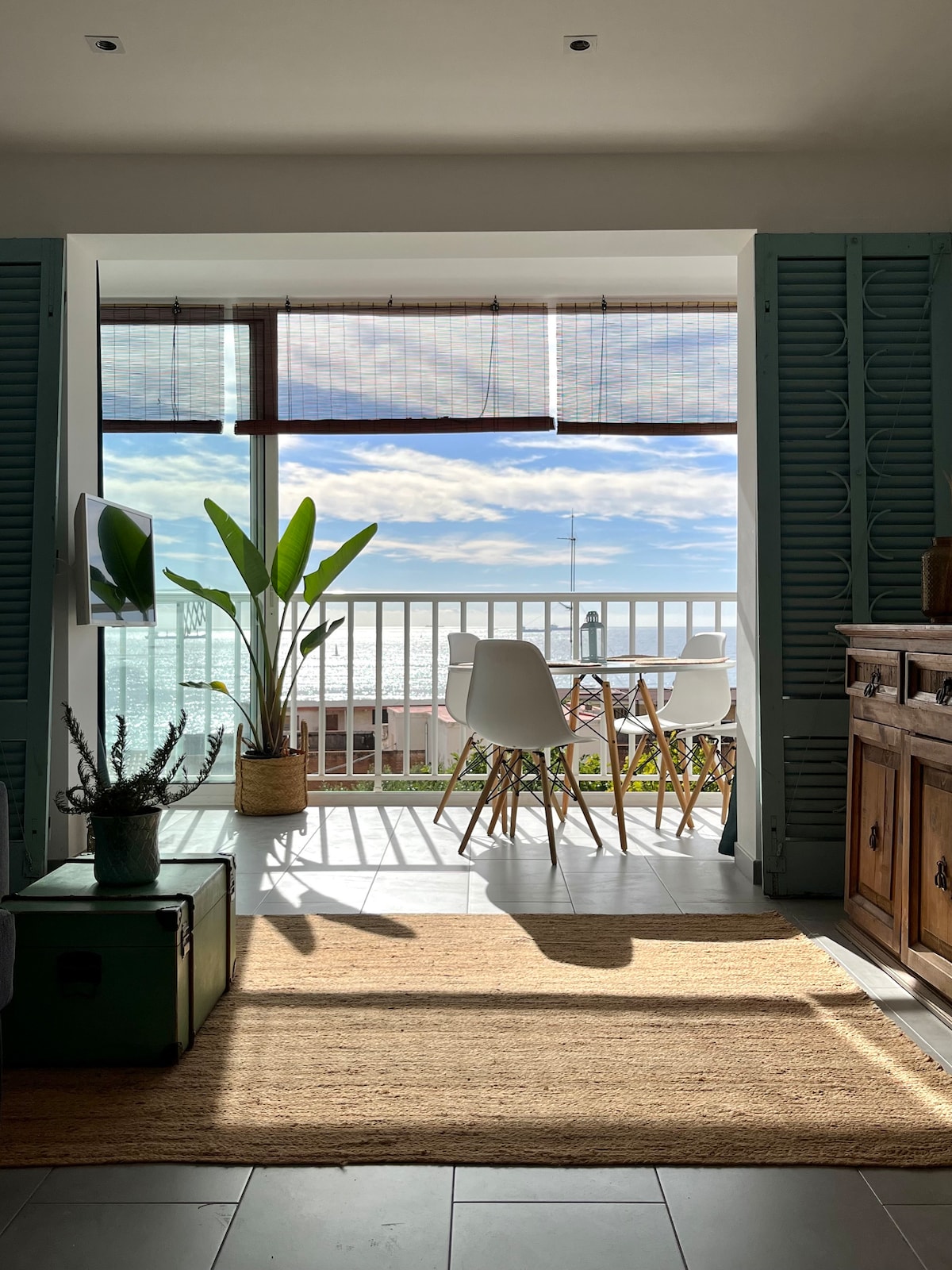 A serene view is captured from an apartment, showcasing a balcony with two modern chairs and a table. Natural light fills the space, highlighting a green plant and warm wooden accents. The ocean is visible beyond the balcony, creating a peaceful ambiance.