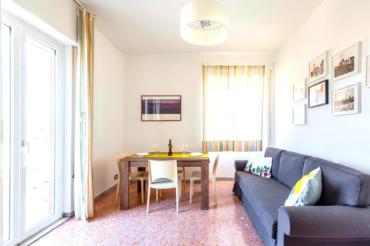 A living area is presented with a beige sofa and a wooden dining table set for four. Large glass doors allow natural light to illuminate the space, complemented by soft window treatments. Wall art adds subtle interest to the neutral walls.