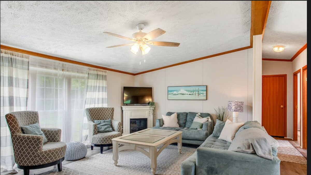 A welcoming living area features a comfortable seating arrangement with two patterned armchairs and a plush sofa. A coffee table sits in the center, while a ceiling fan provides gentle airflow. Natural light fills the space through large windows, complemented by light-colored curtains.