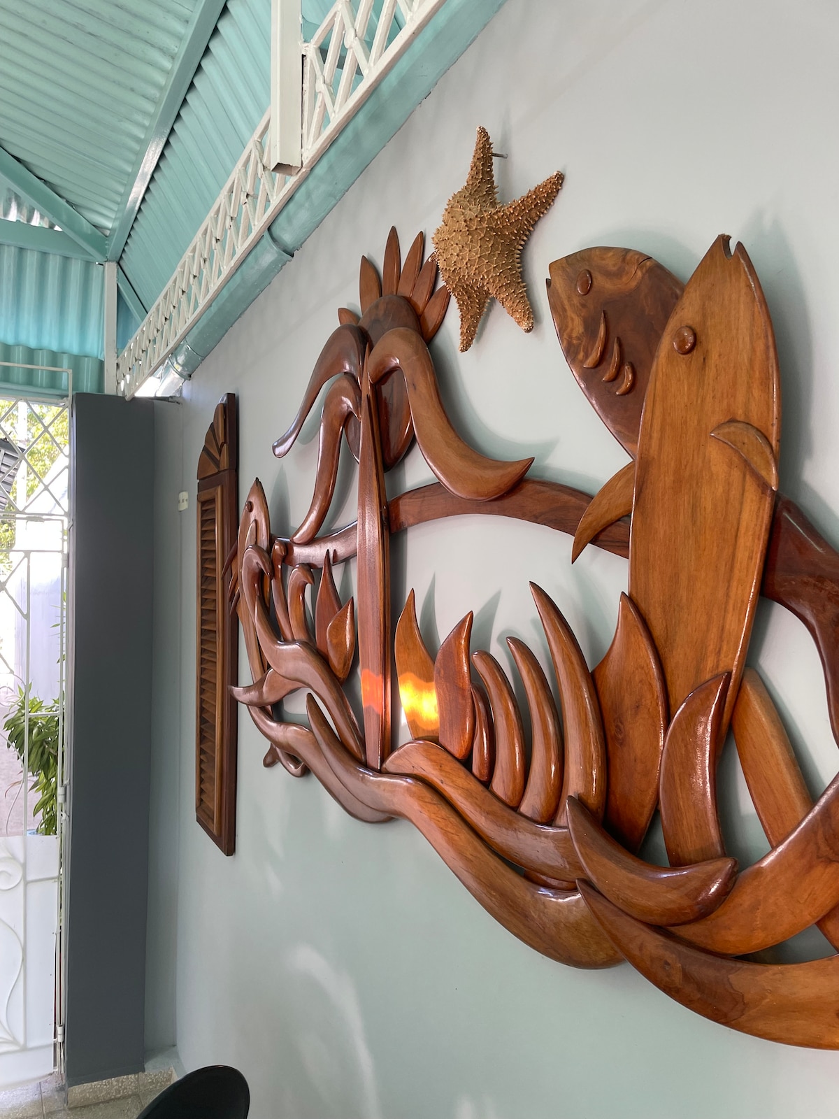 A carved wooden mural depicting fish and sea plants is mounted on a light blue wall. The intricate design includes a starfish, adding a touch of coastal charm to the space. Natural light filters in from a nearby window, enhancing the overall ambiance.