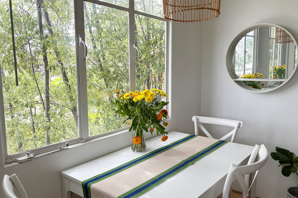 A dining area features a white table set with a floral centerpiece of bright yellow flowers. Natural light filters in through large windows adorned with soft greenery, while a round mirror reflects the serene view. Light-colored chairs complement the inviting setting.