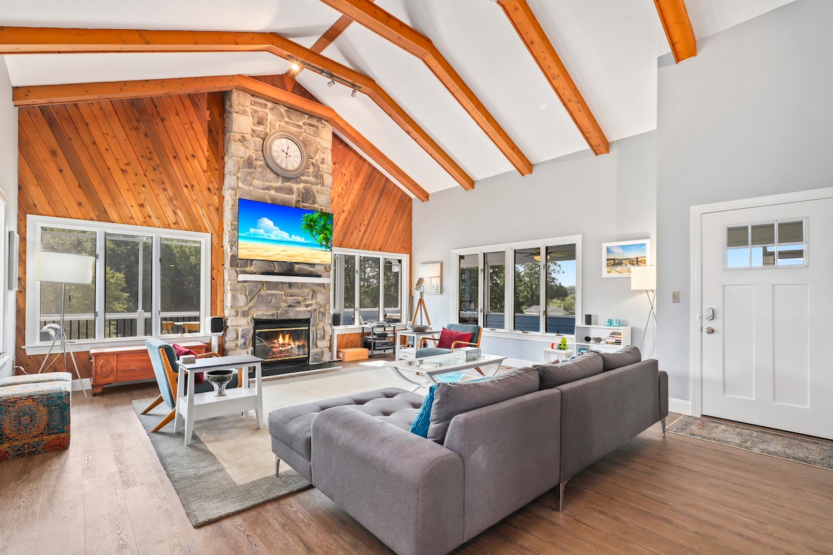 An airy living space features high vaulted ceilings with exposed wooden beams. A stone fireplace serves as a focal point, complemented by a large wall-mounted television. Natural light floods in through expansive windows, illuminating a comfortable seating area that includes a sectional sofa and accent furniture.