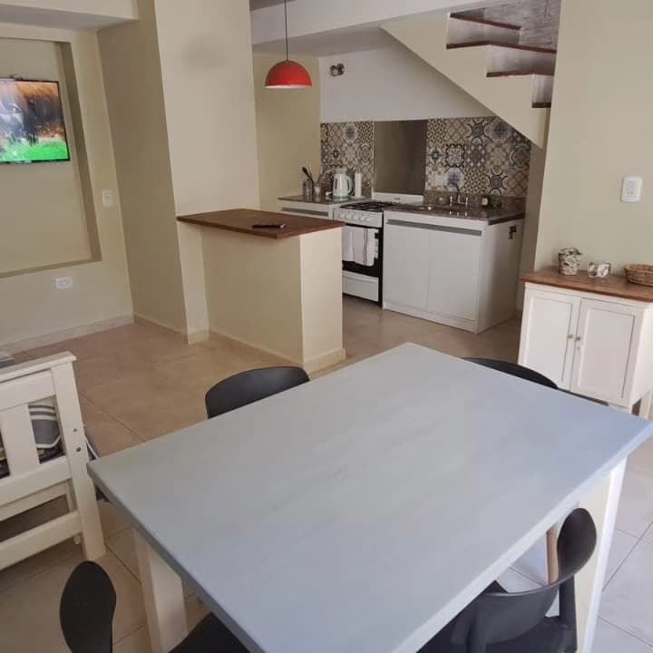 A bright dining area is shown with a light-colored table surrounded by black chairs. An open kitchen is visible in the background, featuring modern appliances and patterned tiles. A staircase leads to the upper level, and a television hangs on the wall.