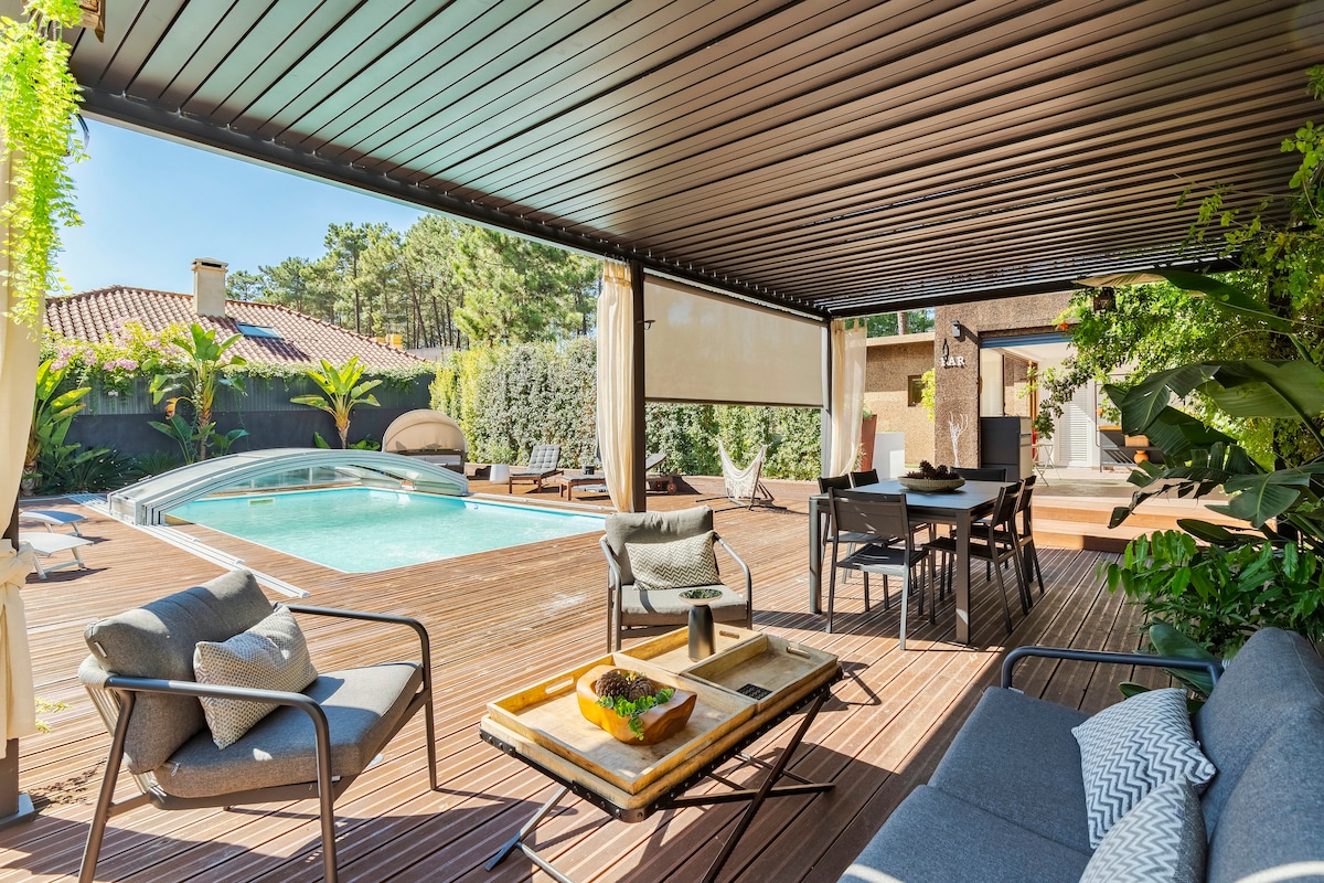A covered outdoor area features comfortable seating arrangements with a small table. A well-maintained pool is visible in the background, surrounded by lush greenery and wooden decking, creating an inviting space for relaxation and socializing.