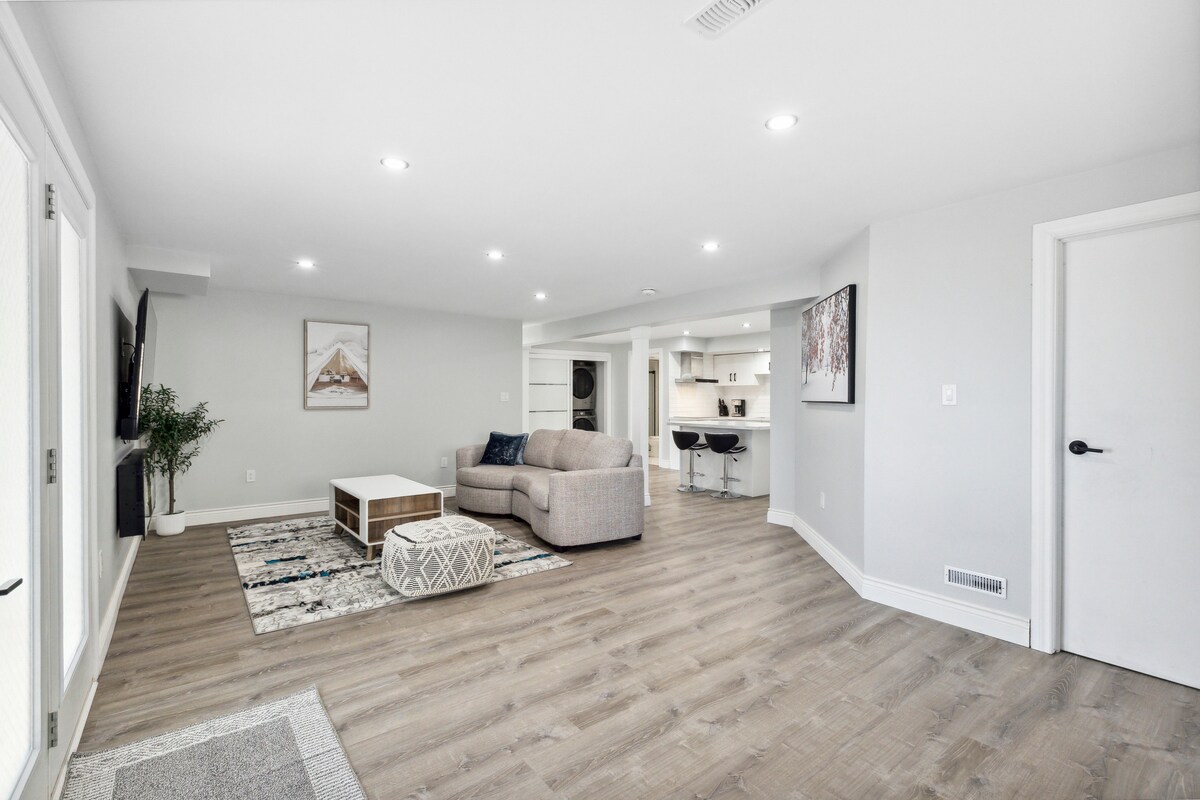 A spacious living area features light hardwood flooring and a neutral color palette. A comfortable sofa is positioned next to a modern coffee table, with a large wall-mounted TV visible. The area connects to a fully equipped kitchen, providing an open and inviting layout.