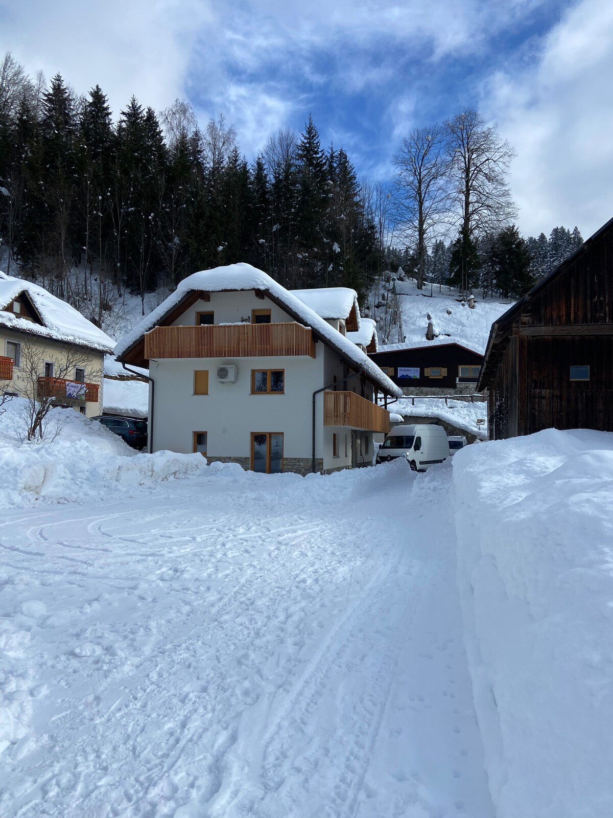 A modern, two-story apartment is nestled in a winter landscape, surrounded by snow-covered trees and mountains. The building features a blend of wood and white exterior, with a sloped roof and large windows. Snow blankets the pathway leading to the entrance.