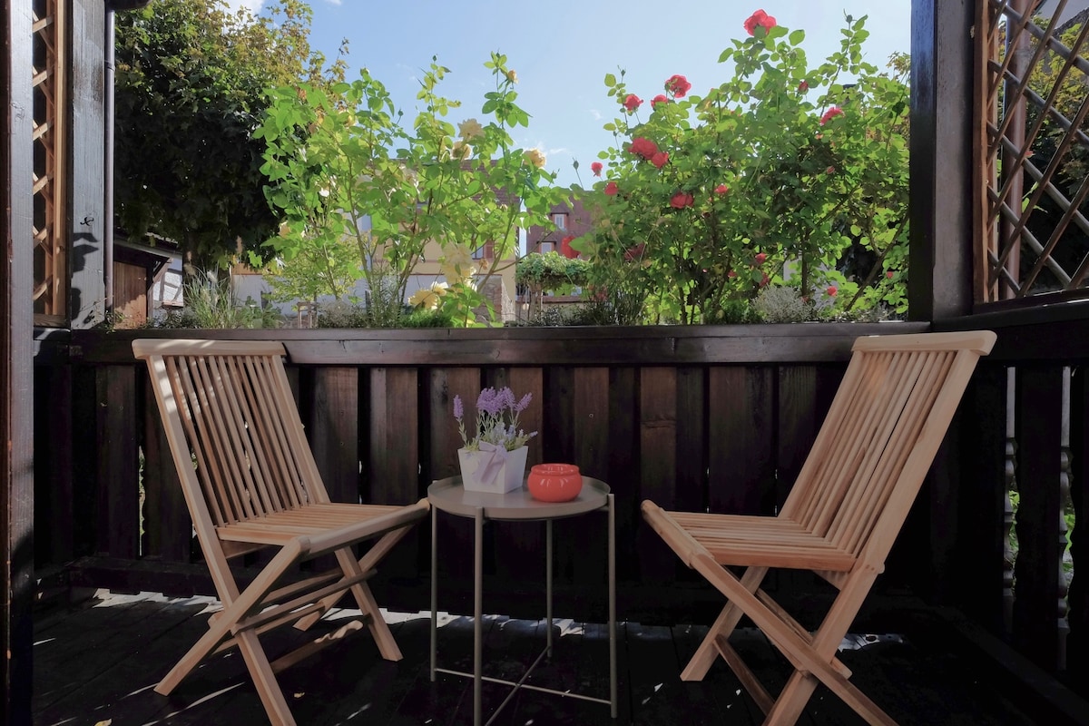 A cozy balcony is depicted, featuring two wooden folding chairs positioned beside a small round table. Bright green foliage and colorful flowers create a natural backdrop, enhancing the outdoor ambiance. Soft sunlight filters through the plants, casting gentle shadows across the space.