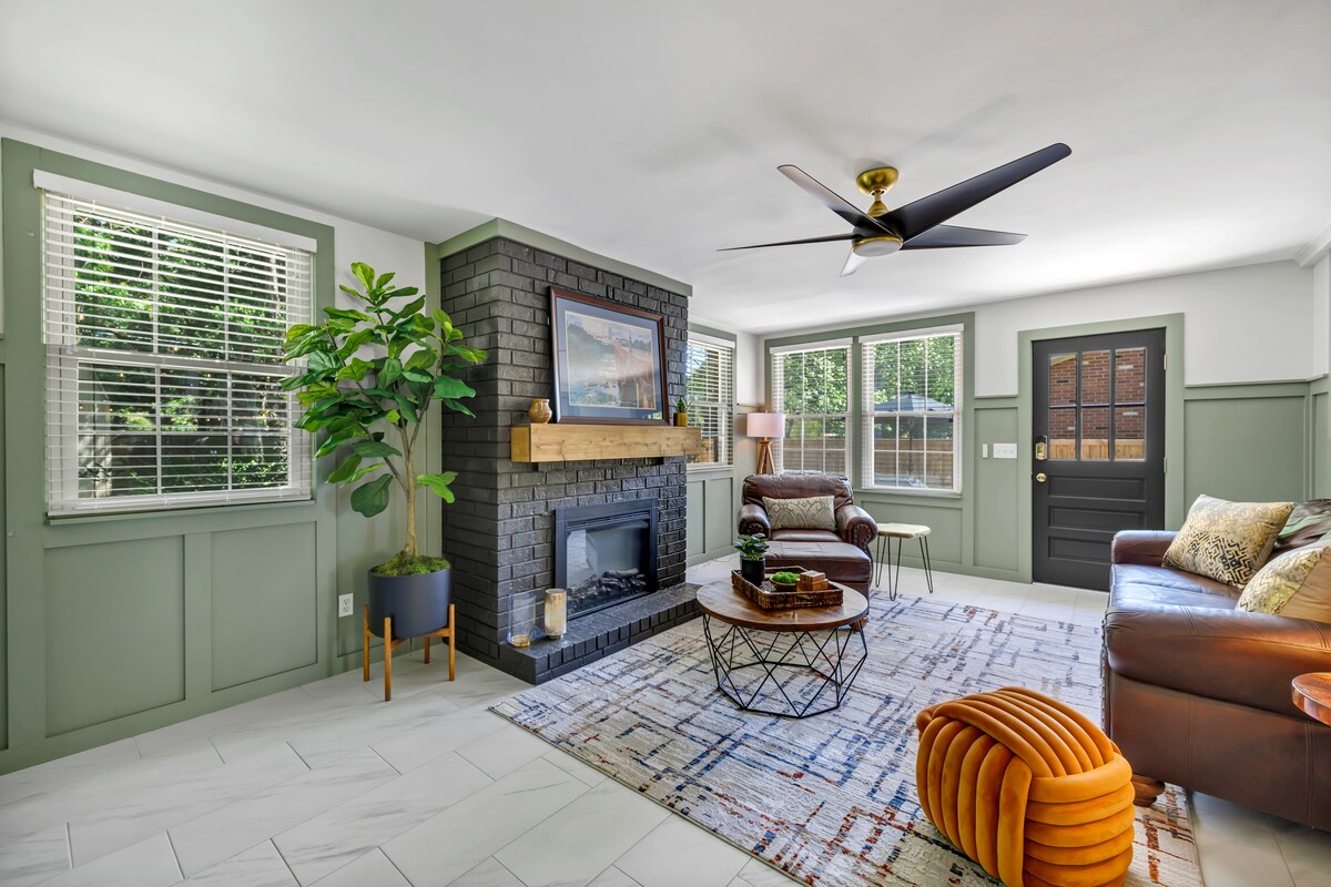 A modern living area features a comfortable leather sofa and a stylish coffee table on a patterned rug. A black fireplace adds an element of warmth, while a ceiling fan enhances airflow. Natural light fills the space through multiple windows, complemented by greenery from a potted plant.