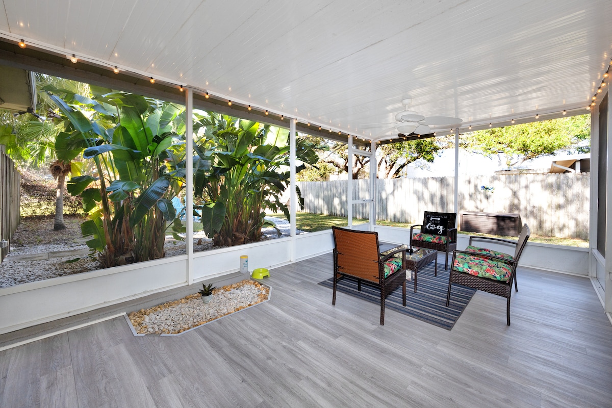 A screened-in patio offers an inviting space with comfortable seating arrangements. Lush greenery is visible through the screens, while a small gravel area adds a natural element. String lights provide a soft glow, enhancing the ambiance for relaxing evenings.