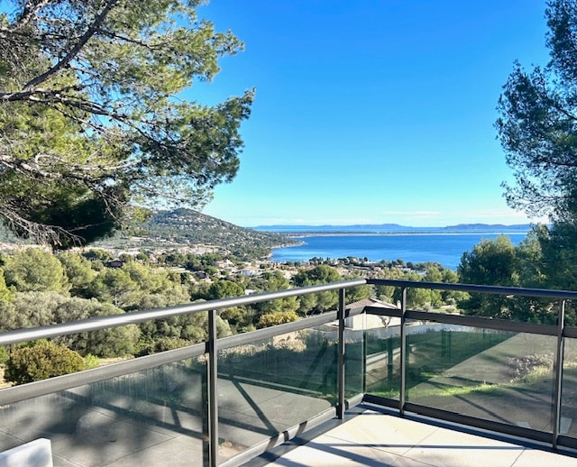 A spacious terrace overlooks the sea and the coastline of Carqueiranne, framed by trees. The view captures the Presqu'Ile de Giens and emphasizes the surrounding natural landscape. The clear sky enhances the serene atmosphere of this outdoor space.