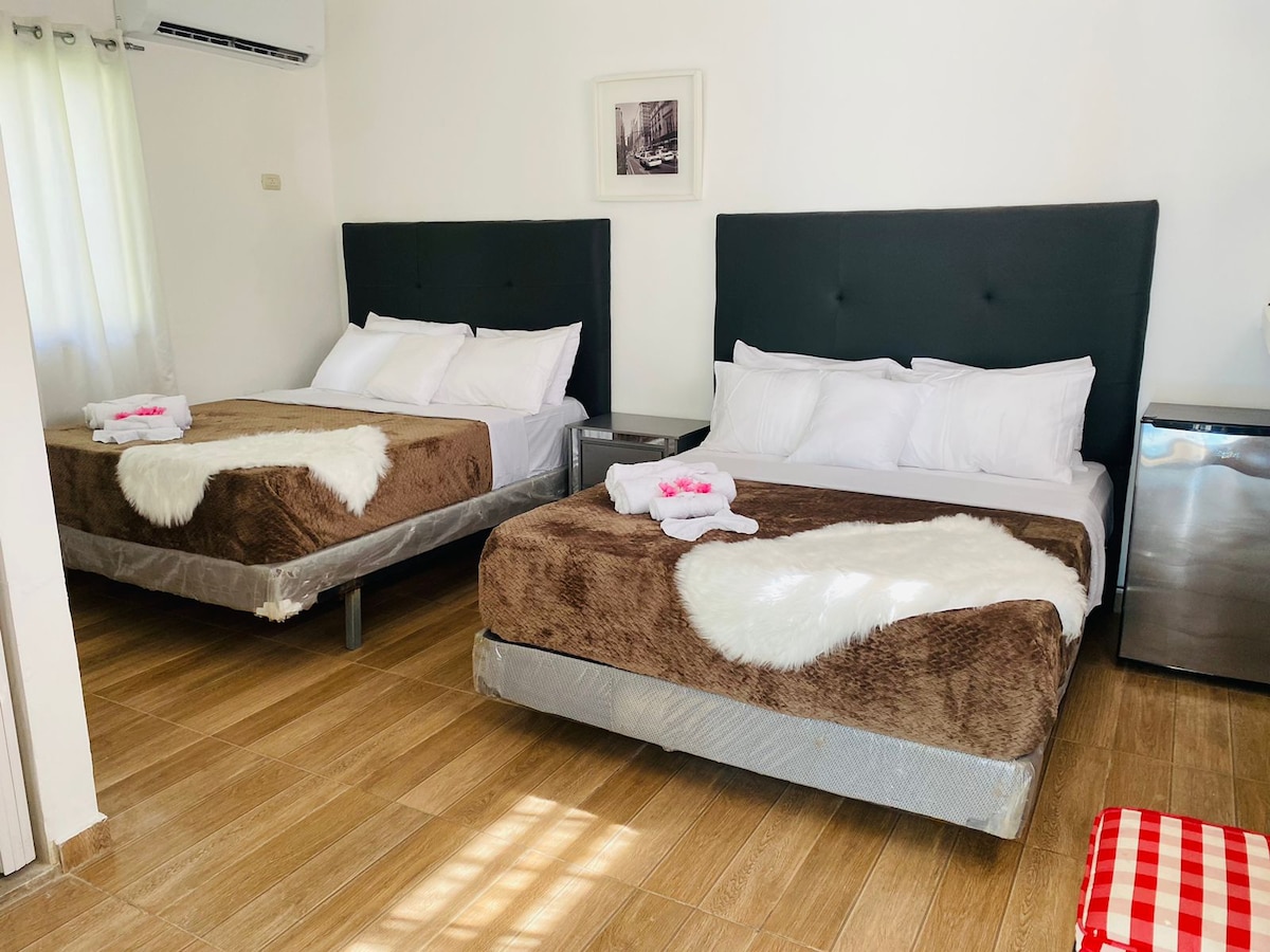 A spacious bedroom features two comfortable beds, each adorned with soft blankets. Light-colored walls and warm wooden flooring create a cozy ambiance. A small modern fridge is situated near the beds, while a framed art piece adds a touch of decor above one of the beds.