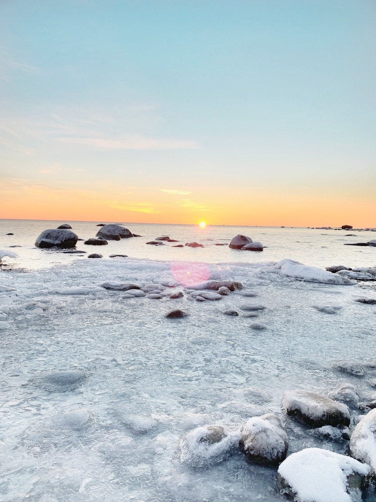 A serene coastal scene is depicted, showcasing a partially frozen shoreline at sunset. Soft hues of orange and pink fill the sky above the calm water. Smooth boulders are scattered along the icy surface, creating an inviting natural landscape.