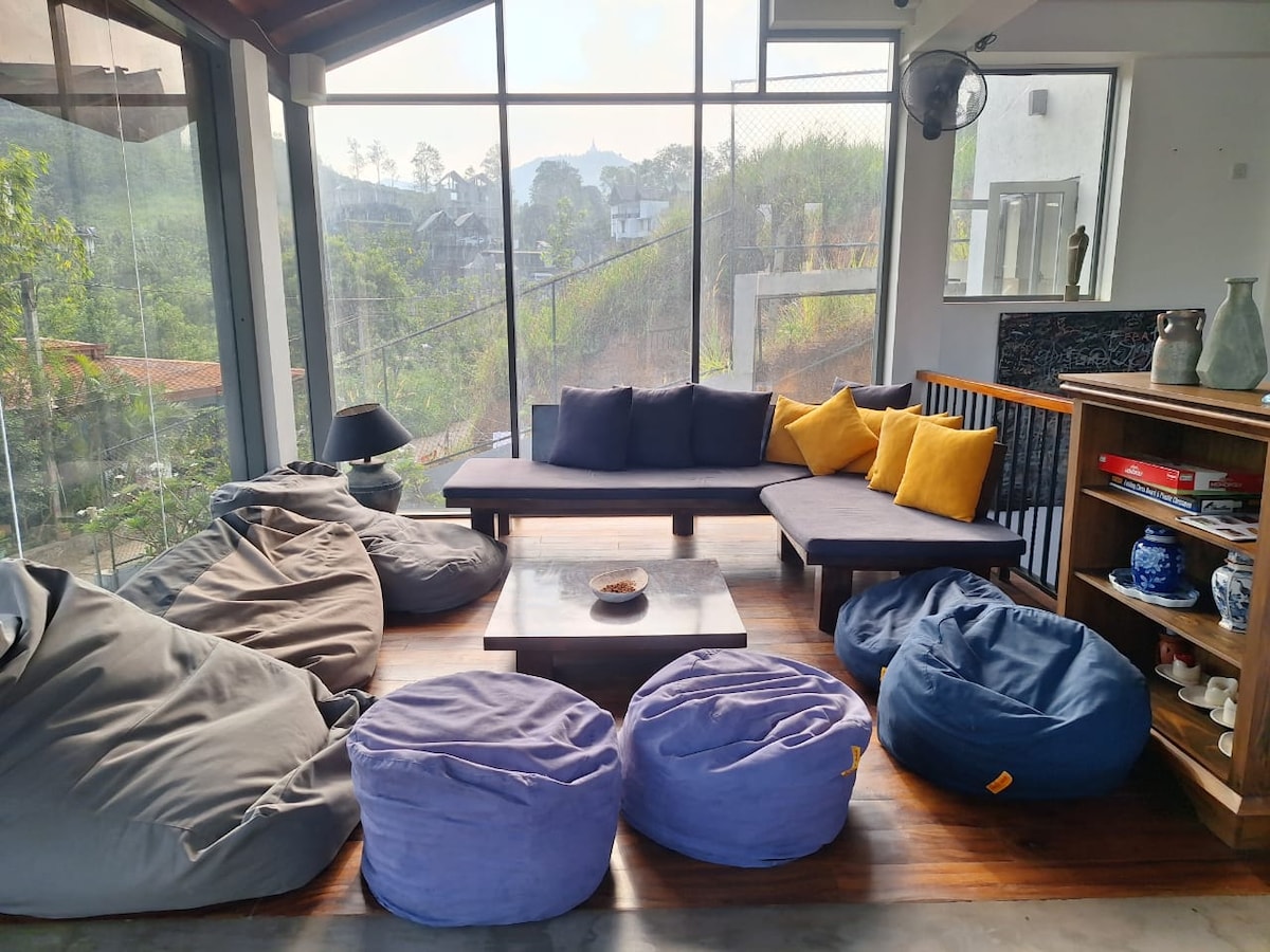 An inviting common area features a large, plush sofa adorned with bright yellow cushions. Several bean bag chairs in soft hues surround a low coffee table. Floor-to-ceiling windows allow natural light to fill the space, providing views of the surrounding greenery.