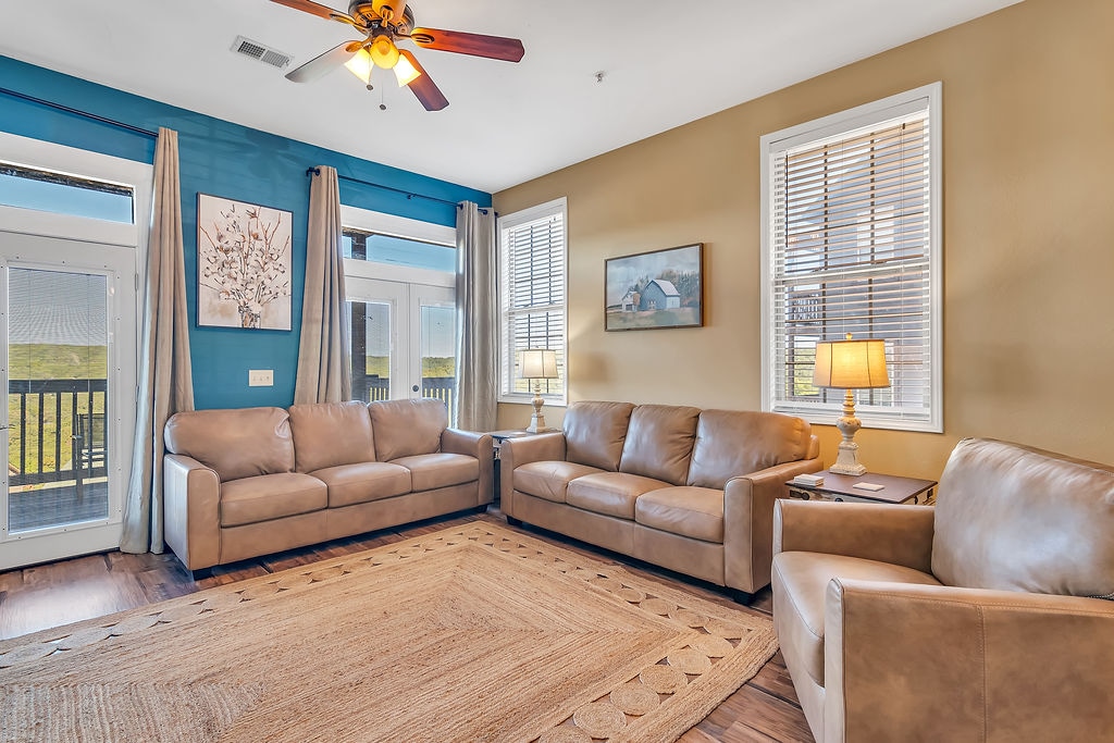 The living room features two beige leather couches and a single armchair, arranged around a large area rug. Natural light streams through two windows, and a sliding glass door opens to an outdoor balcony. A ceiling fan provides additional comfort.