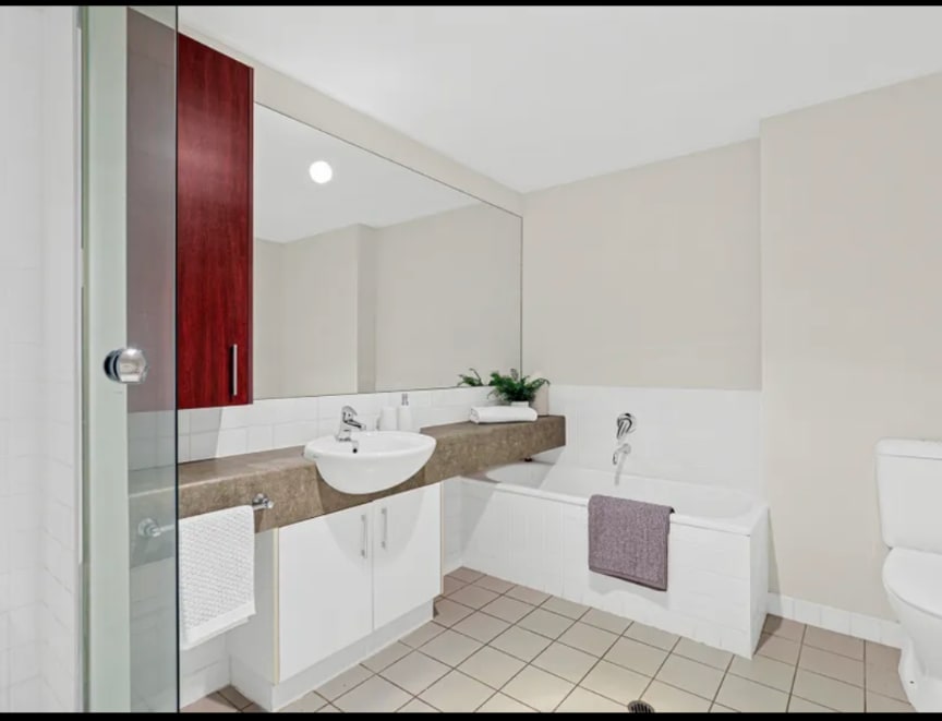 A well-appointed bathroom features a spacious vanity with a large mirror and a sink. A modern tub is situated beside the shower, adorned with a gray washcloth. Light beige tiles create a neutral backdrop, complemented by wooden cabinetry and a potted plant.