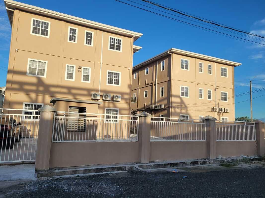 A two-building complex is seen enclosed by a short fence. Each structure features multiple windows with air conditioning units below them, providing a sense of modern functionality. The clear blue sky enhances the neutral tones of the buildings, creating a welcoming appearance.