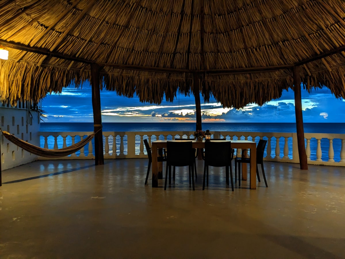 A covered outdoor dining area is enclosed by a thatched roof, with a large table and several chairs positioned for ocean views. The horizon displays a gradient of colors as the sun sets over the calm water, creating a serene backdrop.