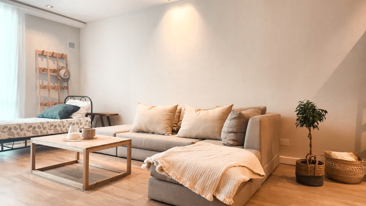 The bright living area features a comfortable sectional sofa and a light wooden coffee table. A cozy throw blanket is casually placed on the sofa. A separate bed is also visible in the room, accompanied by a small plant and a woven basket.