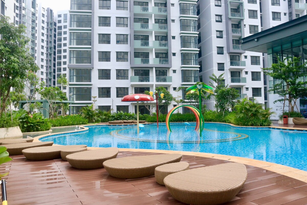 A large pool is surrounded by lush greenery and modern buildings. Curved lounge chairs made of wicker are positioned around the pool area. Colorful water play structures are visible in the shallow end, creating an inviting space for relaxation and leisure.