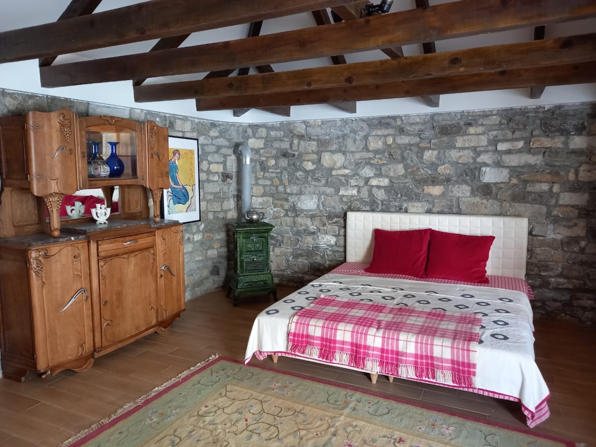 A cozy sleeping area features a spacious bed adorned with red pillows and a patterned blanket. A wooden cabinet adds character against the stone wall, complemented by decorative items. Exposed wooden beams highlight the room's rustic charm, while a patterned rug adds warmth to the floor.