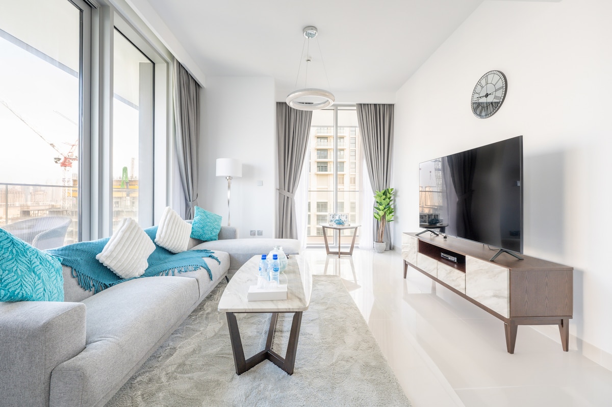 A spacious living area features large floor-to-ceiling windows, allowing ample natural light. A grey sofa adorned with turquoise cushions is positioned beside a low coffee table. A flat-screen television is mounted on a sleek cabinet, while decorative plants add greenery to the space.