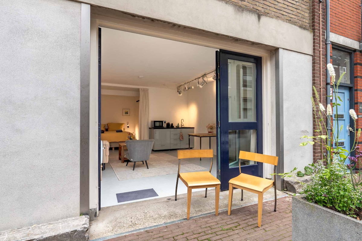 A modern entrance features two wooden chairs positioned outside the spacious open doorway. Inside, a well-lit living area showcases a seating arrangement and a kitchenette, all designed with neutral tones. Fresh greenery is visible in nearby planters, enhancing the urban environment.