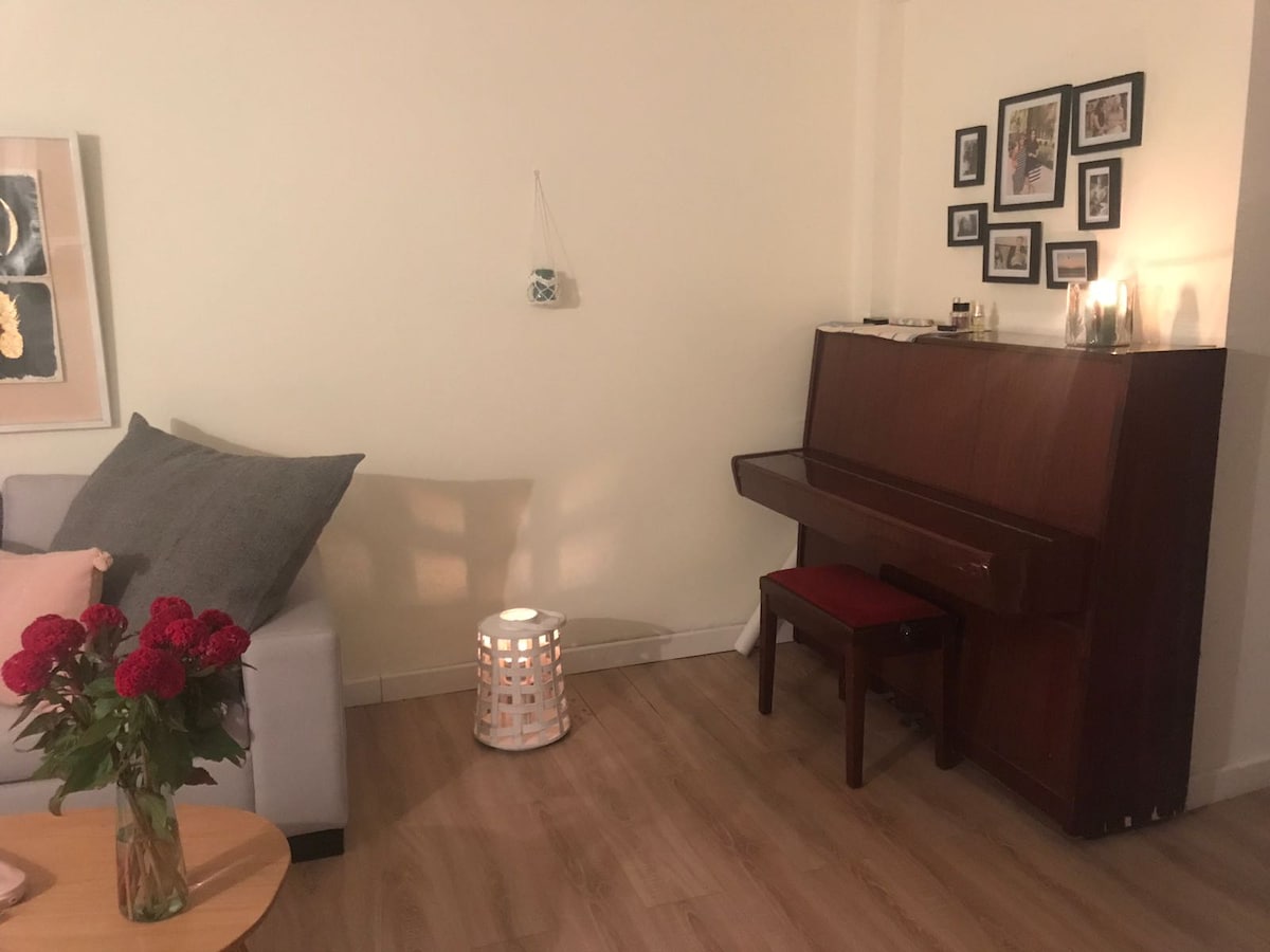 A warm living space features a light-colored sofa with decorative pillows. A wooden piano stands against the wall, accompanied by a small red stool. A vase of roses is displayed on a round table, while soft lighting emits from a nearby lantern.