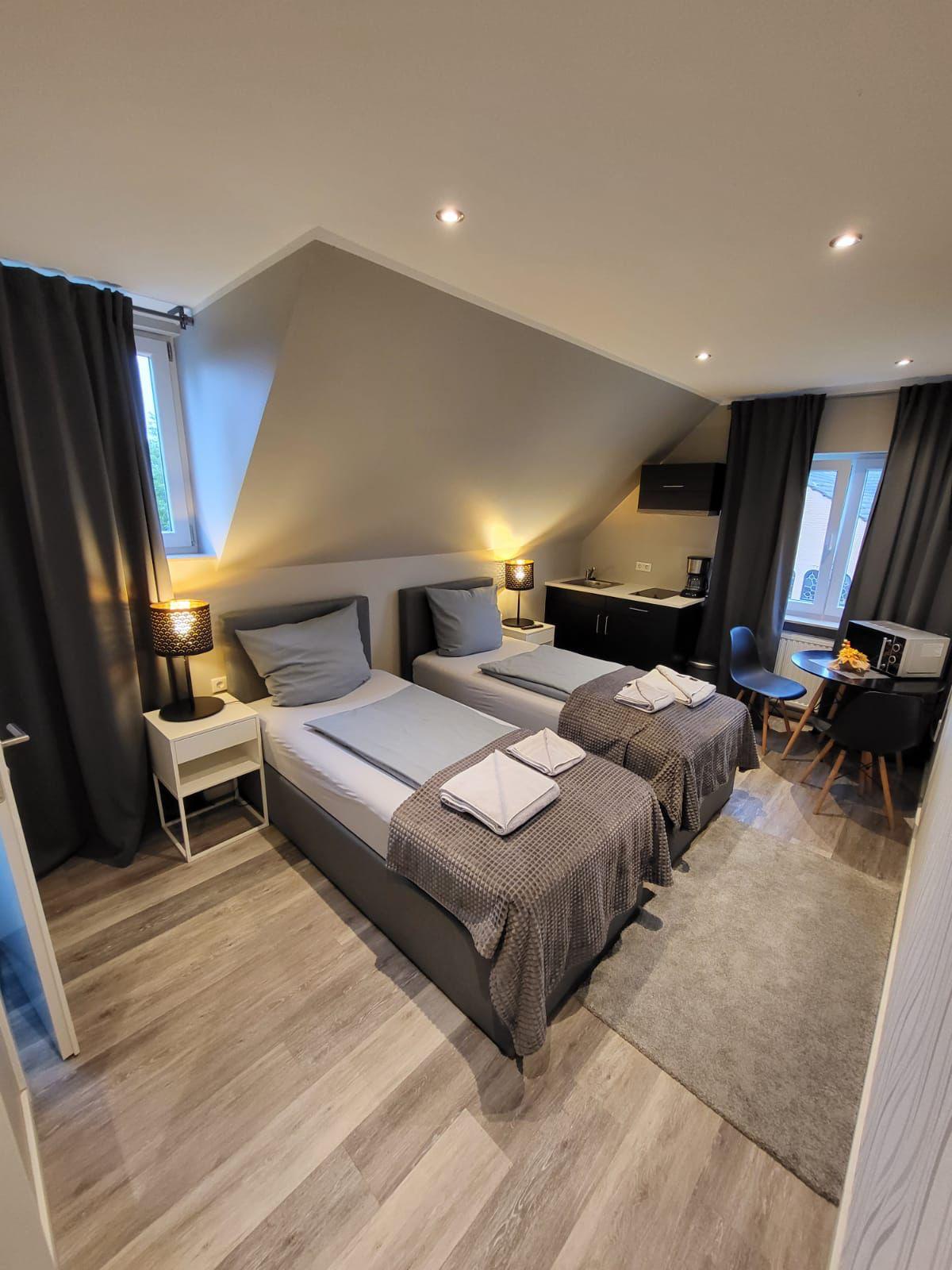 A well-lit room features two twin beds neatly arranged with gray linens and towels. Dark curtains frame a window, allowing natural light to brighten the space. A small dining area with a table and chairs is present, accompanied by cabinetry and a microwave.