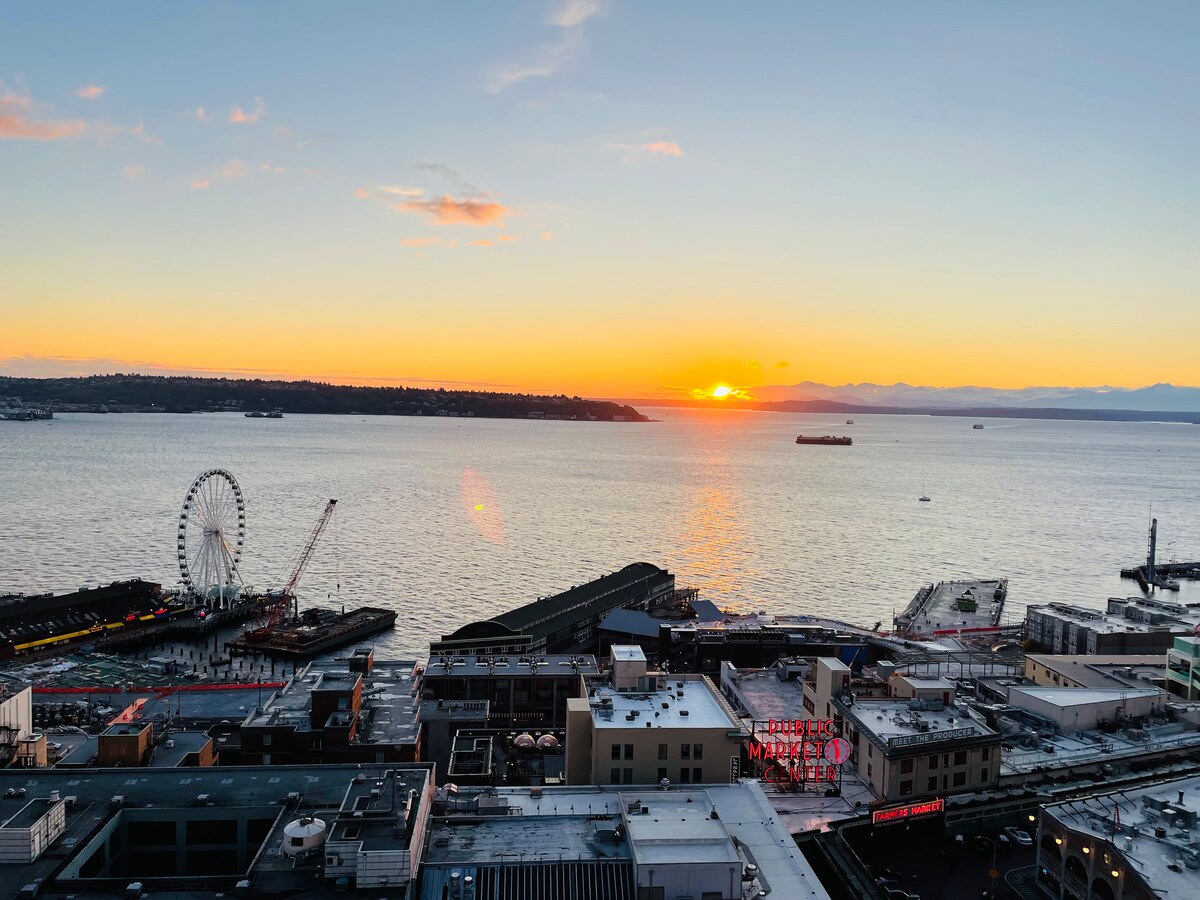 A scenic view of the Puget Sound at sunset, showcasing the vibrant colors of the sky reflected on the water. The Seattle Great Wheel and nearby structures are visible along the waterfront. The distant Olympic Mountains create a striking backdrop, enhancing the natural beauty of the scene.