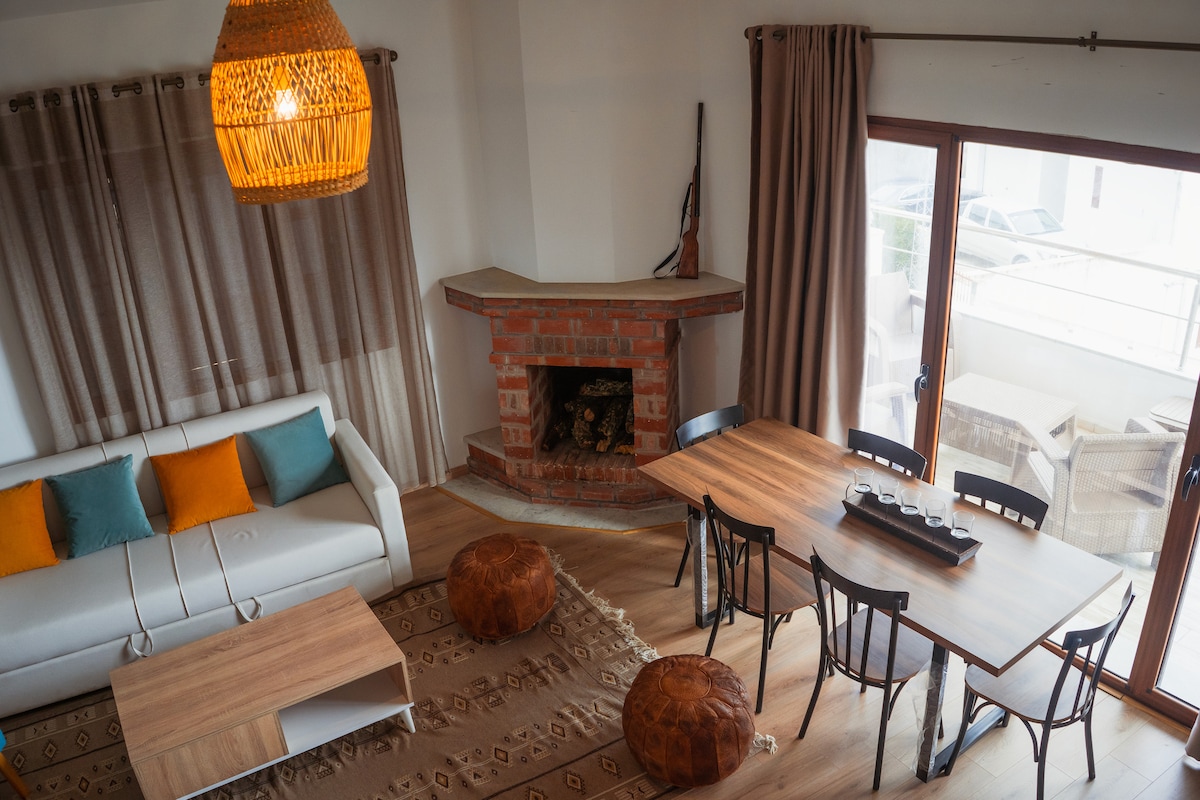 A cozy living area features a white sofa adorned with teal and orange cushions. A wooden coffee table sits on a decorative rug, while a dining table with black chairs is positioned nearby. A brick fireplace adds warmth to the space, complemented by softly draped curtains.