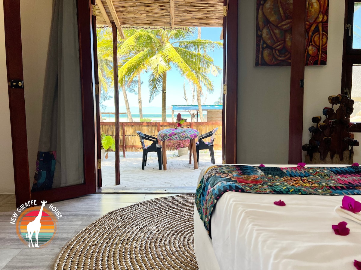 An inviting room with a spacious bed is viewed through open doors, leading to a sandy outdoor area. A small table with chairs overlooks the beach, framed by swaying palm trees and the ocean in the background.