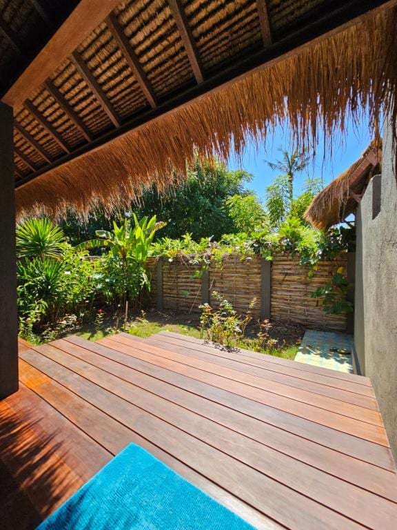 A wooden deck is presented in the foreground, leading to a lush garden area surrounded by greenery. Sunlight filters through the leaves, creating a serene outdoor space. Thatch roofing is visible overhead, adding to the tropical ambiance.