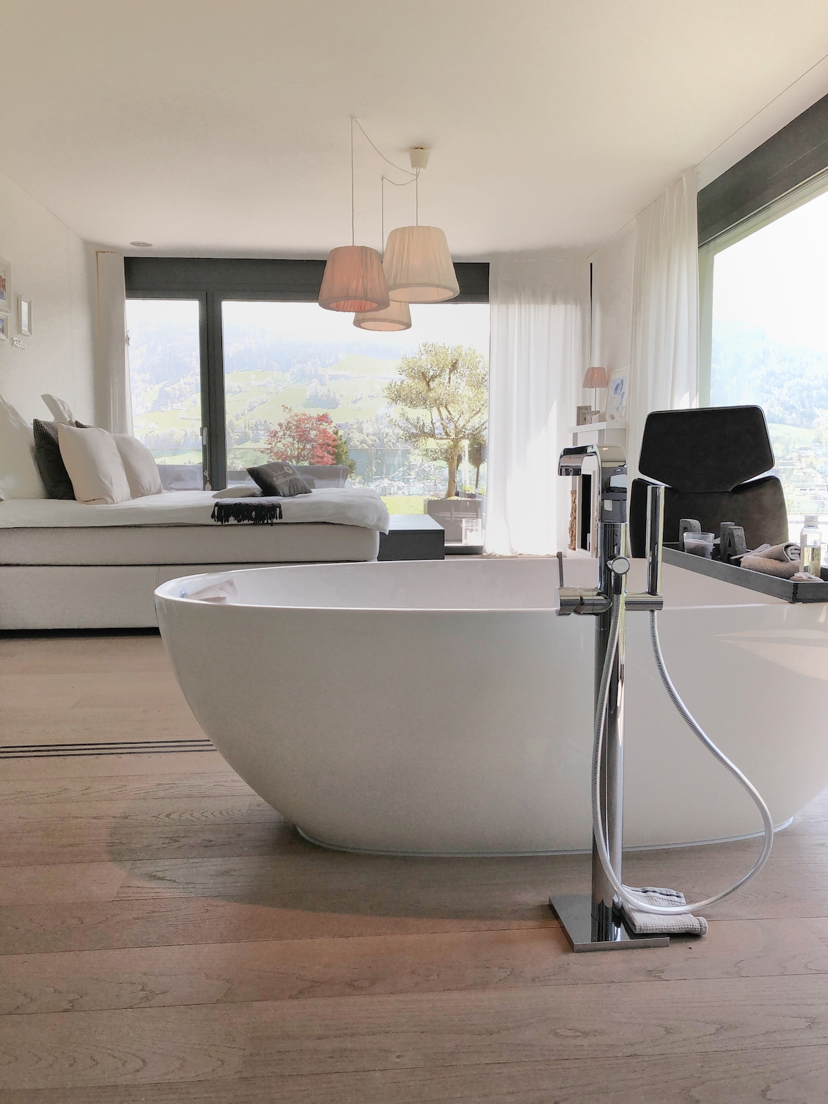 An elegant bathroom space features a freestanding soaking tub in a contemporary design. A large window offers views of the surrounding landscape, while a cozy seating area with light-colored furnishings is visible in the background. Natural light enhances the serene atmosphere.