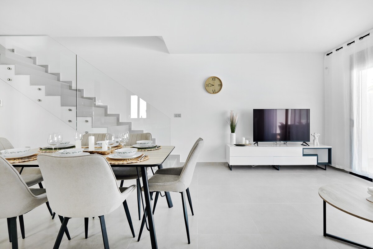 The dining area is furnished with a light-colored table surrounded by six upholstered chairs. A flat-screen TV is positioned against a white media console. Natural light filters through sheer curtains, and a stylish clock adorns the wall, complementing the modern design.