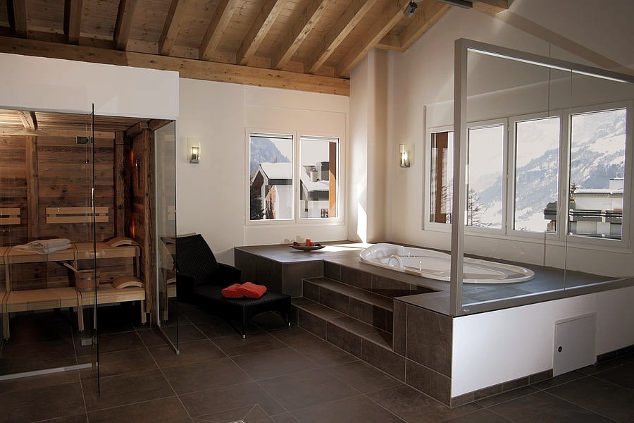 A wellness area features a wooden sauna adjacent to a sleek, modern whirlpool tub. Large windows provide natural light and view of the mountains. A black lounge chair is positioned nearby, accompanied by a small tray with refreshments.