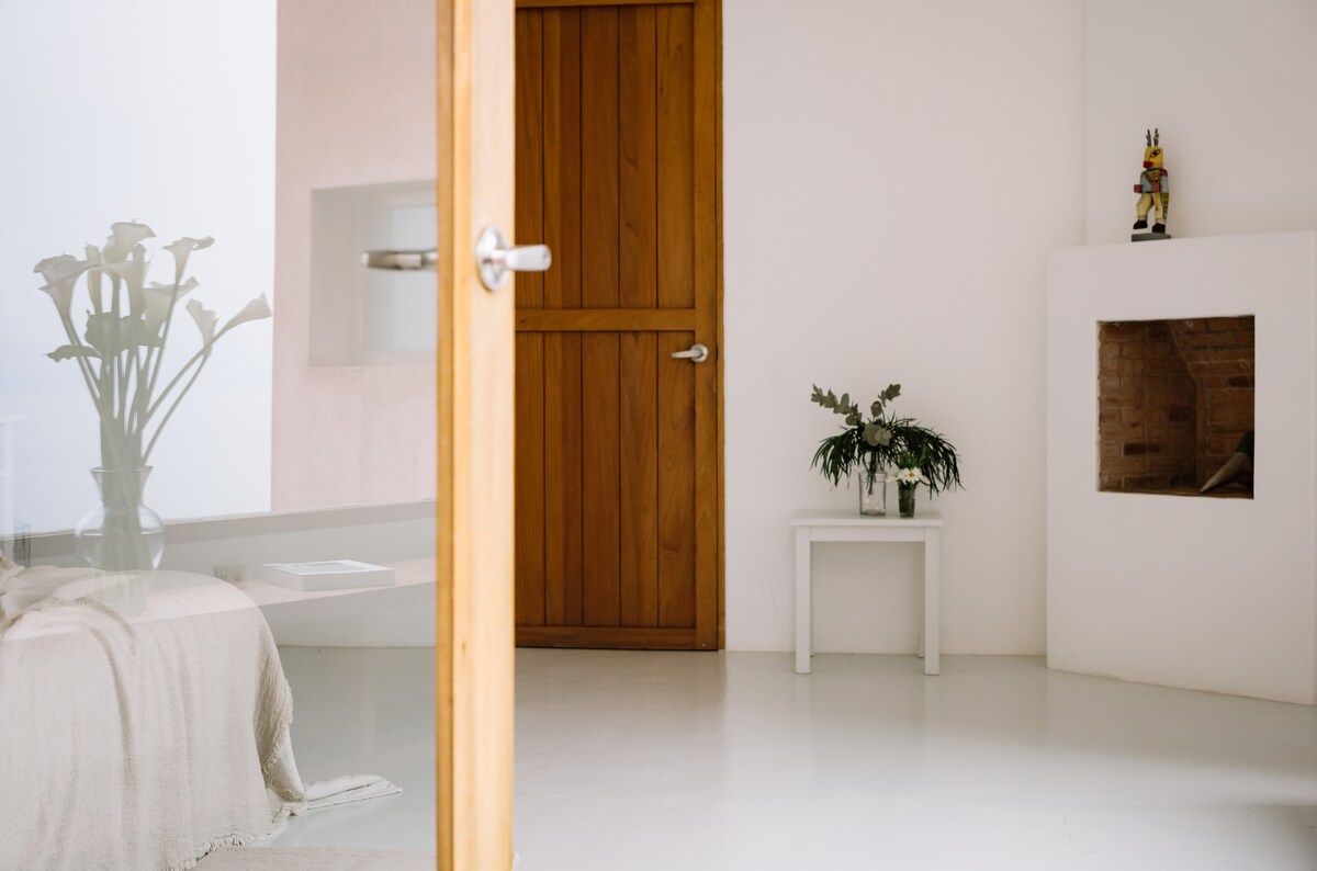 The image captures a serene interior space featuring a wooden door leading to a minimalist room. A small white table with delicate greenery sits beside a fireplace. The clean, bright walls and polished floor enhance the tranquil ambiance.