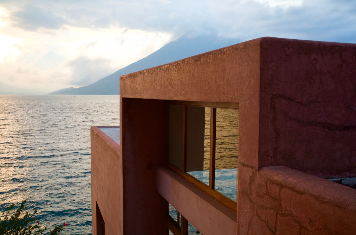A pink architectural structure is seen against the backdrop of a glistening lake, framed by distant mountains. The expansive glass windows invite natural light, creating a serene connection with the surrounding landscape.