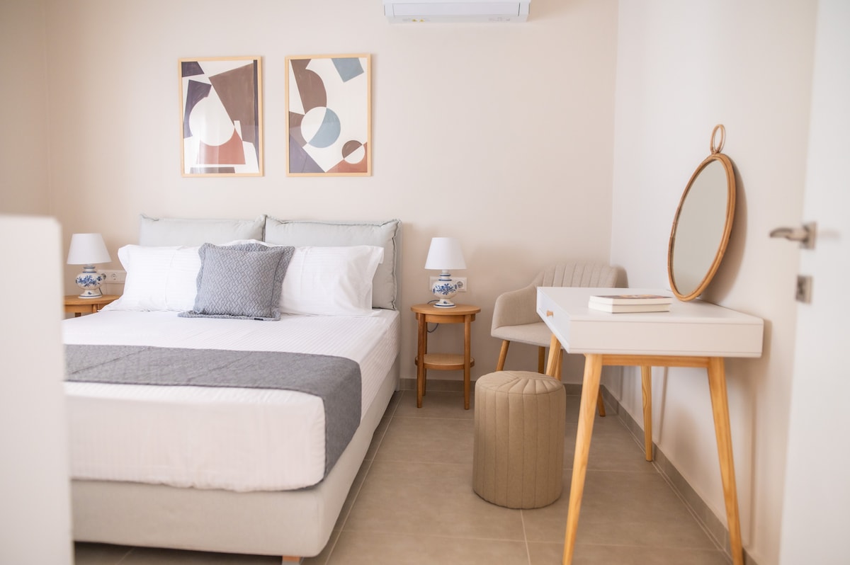 A serene bedroom is presented with a spacious bed adorned with crisp white linens and a soft grey throw. Two framed artworks hang above the bed, while a stylish mirror, a small round stool, and bedside lamps complete the elegant design.