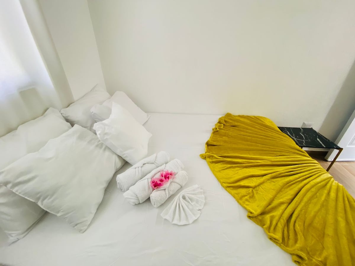 A cozy sleeping area is presented with a spacious bed adorned with an assortment of white cushions. Soft towels are arranged neatly alongside a vibrant yellow blanket draped elegantly over the bed, creating a calming visual appeal.
