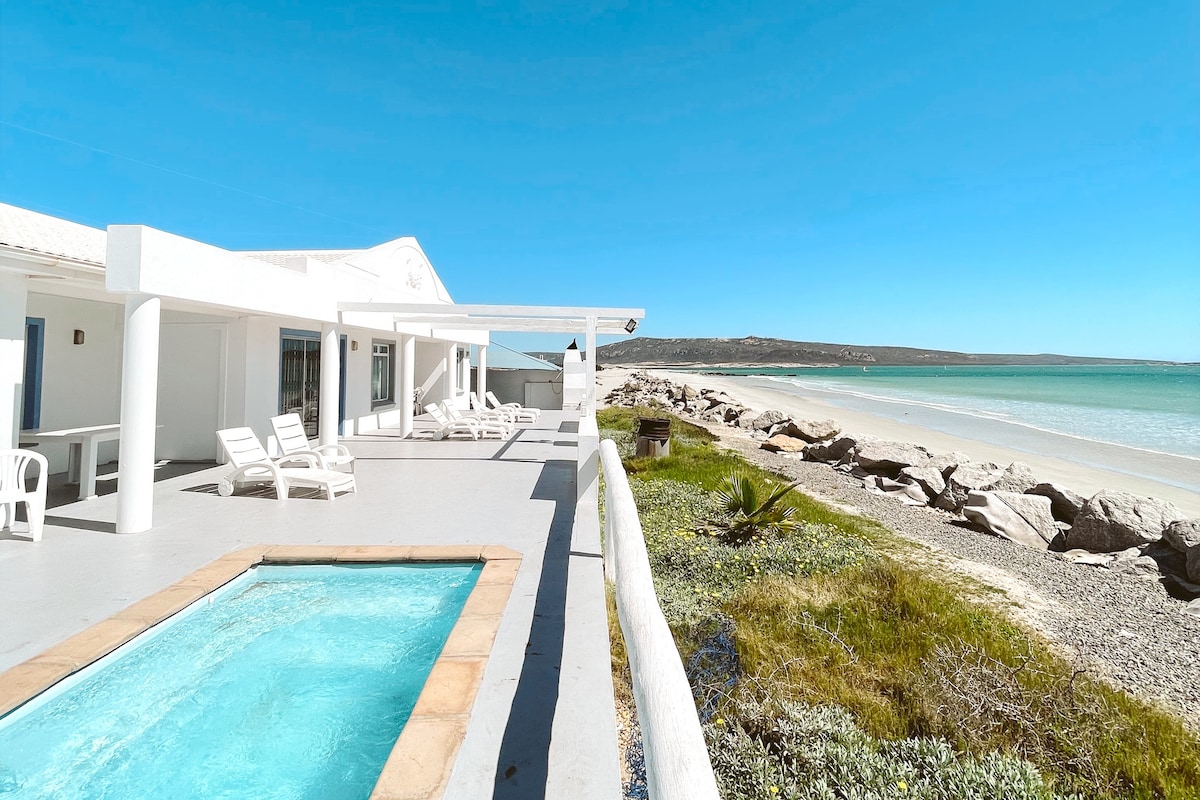 A sunlit terrace is highlighted, featuring lounge chairs positioned around a small pool. A clear view of the sandy beach and turquoise lagoon is visible, with the shoreline extending in the distance. The bright blue sky complements the white architectural details of the property.