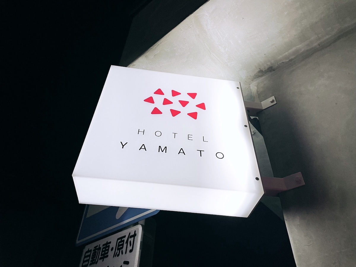 A backlit sign displays the name 'HOTEL YAMATO' in bold letters, framed by a series of red triangle shapes. The sign is positioned above a street, providing clear visibility in low light conditions.