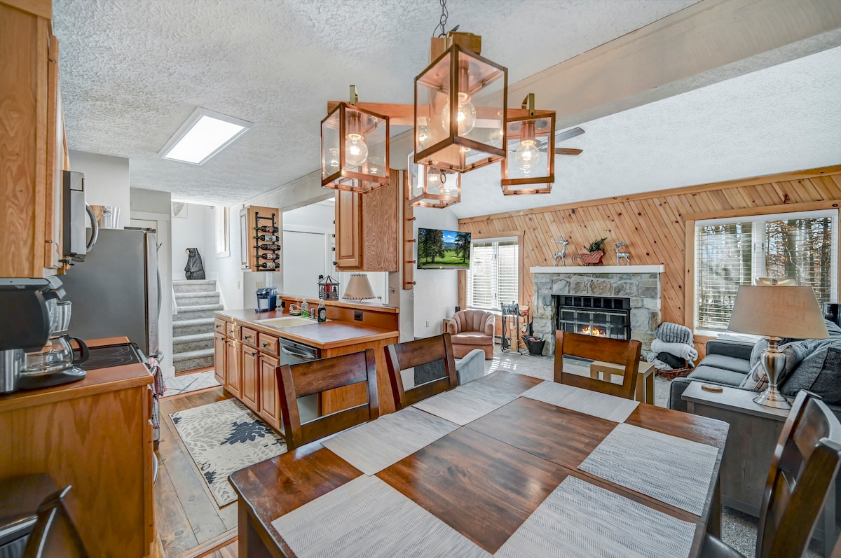 A warm dining area features a large wooden table with an inviting arrangement of light and dark panels. Above, pendant lights hang gracefully, illuminating the space. A cozy living area with a stone fireplace is visible in the background, alongside a wall-mounted television.