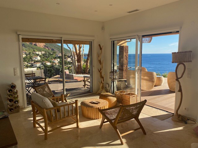 A bright living space is highlighted by large glass doors that open to a stunning sea view. Natural light fills the room, showcasing wooden decor and comfortable furniture. A cozy seating area includes a round table and bamboo chairs, creating a relaxed environment with a coastal ambiance.