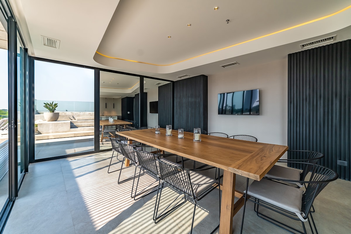 A modern dining area features a large wooden table surrounded by wire chairs, providing seating for multiple guests. Floor-to-ceiling windows allow natural light to illuminate the space, while views of a patio area are visible in the background.