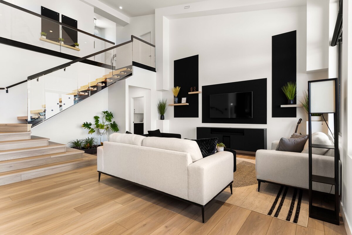 A spacious living area features a large light-colored sofa facing a sleek black entertainment unit. Mixed greenery adds a touch of nature, while wood flooring and a stylish rug create a warm and inviting setting. A modern staircase with glass railings is visible in the background.