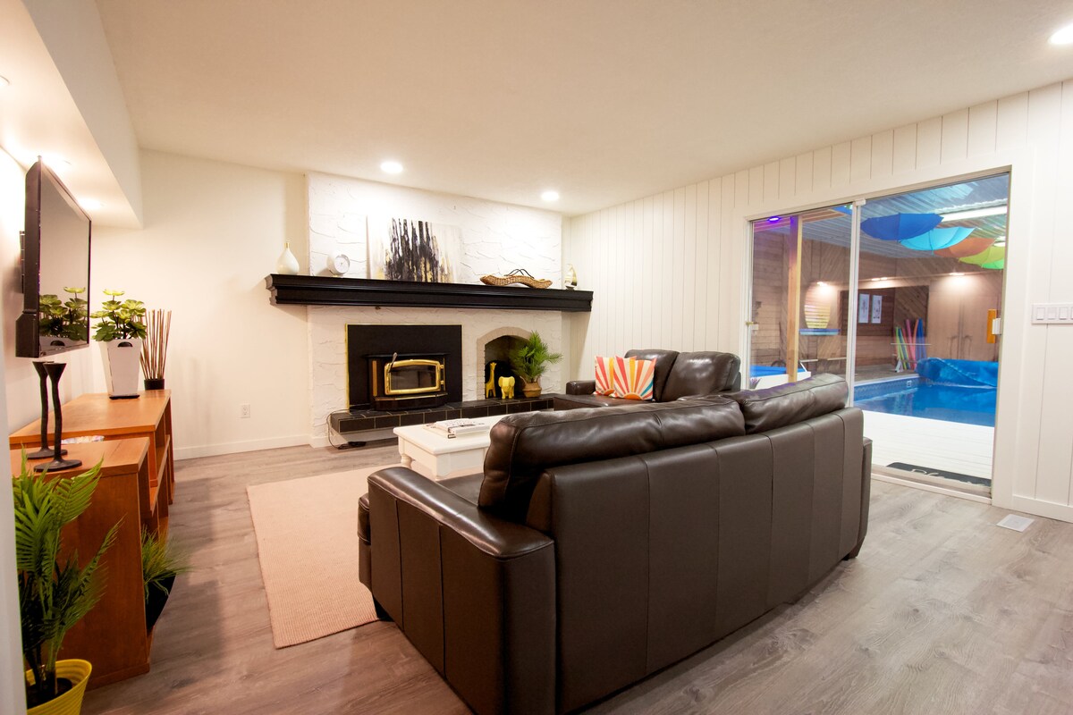 A spacious living area features large windows that provide views of the indoor pool. The seating arrangement includes a comfortable dark leather sofa, complemented by a light-colored area rug. A fireplace creates a cozy focal point, enhancing the welcoming atmosphere.