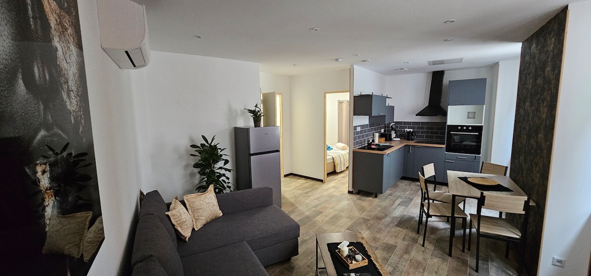 A modern living space features a comfortable grey sofa and sleek dining area. The kitchen is equipped with contemporary appliances, while light-colored cabinetry contrasts with dark accents. Potted plants add a touch of greenery, enhancing the overall ambiance of the room.