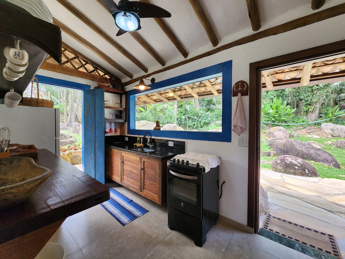 A well-equipped kitchen features wooden cabinetry and a black countertop. Natural light enters through large windows, providing views of the lush outdoors. A stove sits beside a white countertop, and a small rug adds a touch of color to the tiled floor.