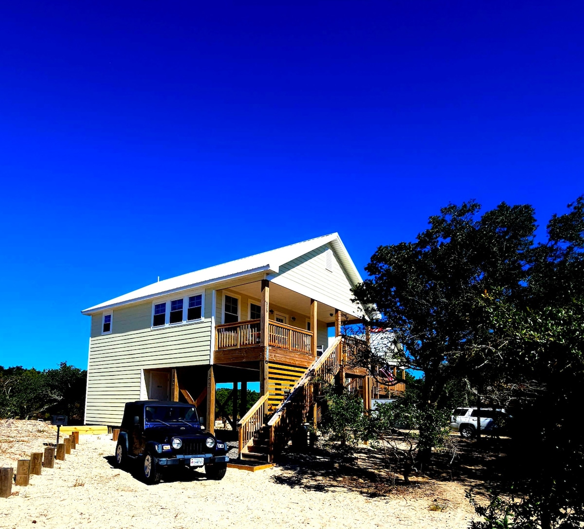 The two-story cottage features a light-colored exterior and a spacious deck with stairs leading to the entrance. Two vehicles are parked in the sandy area, surrounded by natural vegetation and a clear blue sky.