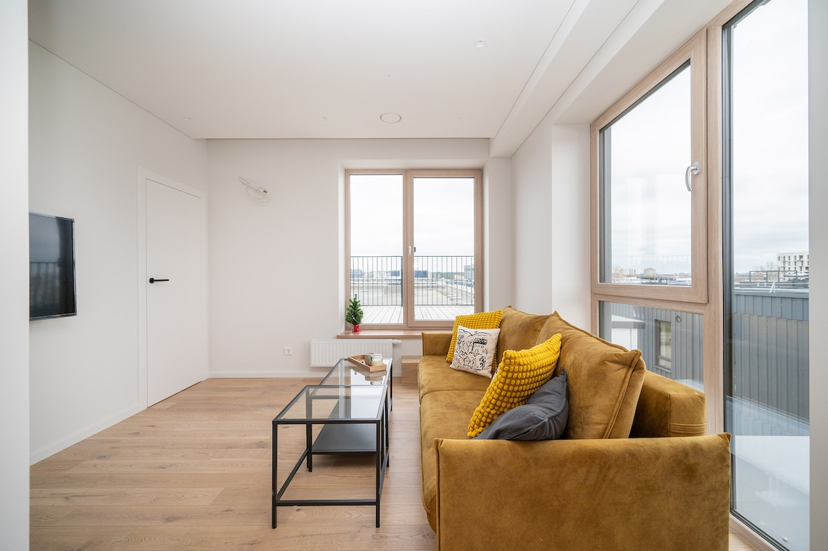 A bright living area features a comfortable sofa in warm tones, complemented by a decorative pillow. A glass coffee table sits in front, with a small potted plant nearby. Large windows provide ample natural light and offer views of the surrounding area.